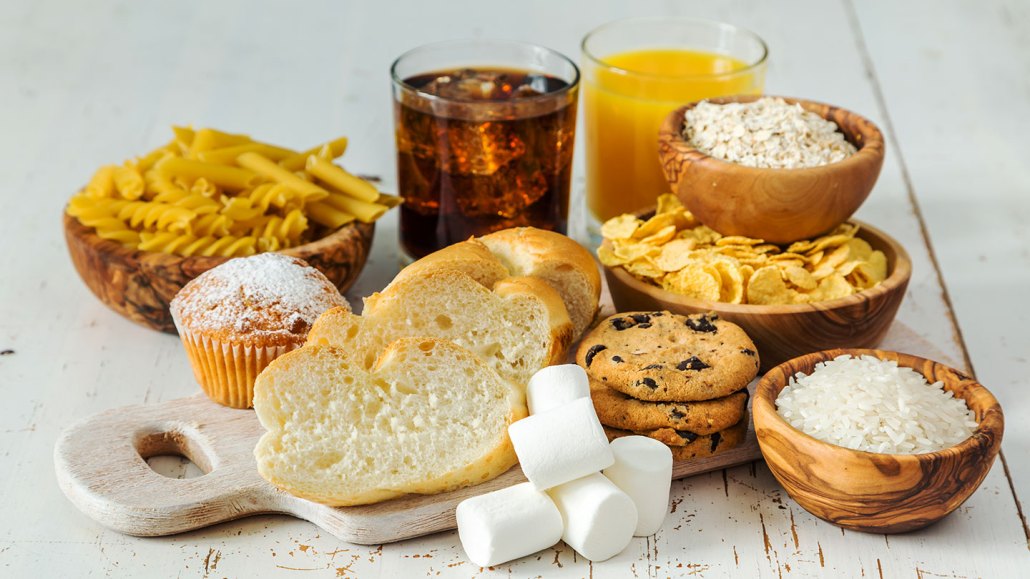 a selection of carbohydrates on a table: sliced bread, pasta, marshmellows, soda and juice, rice, potato chips, chocolate chip cookies and oats