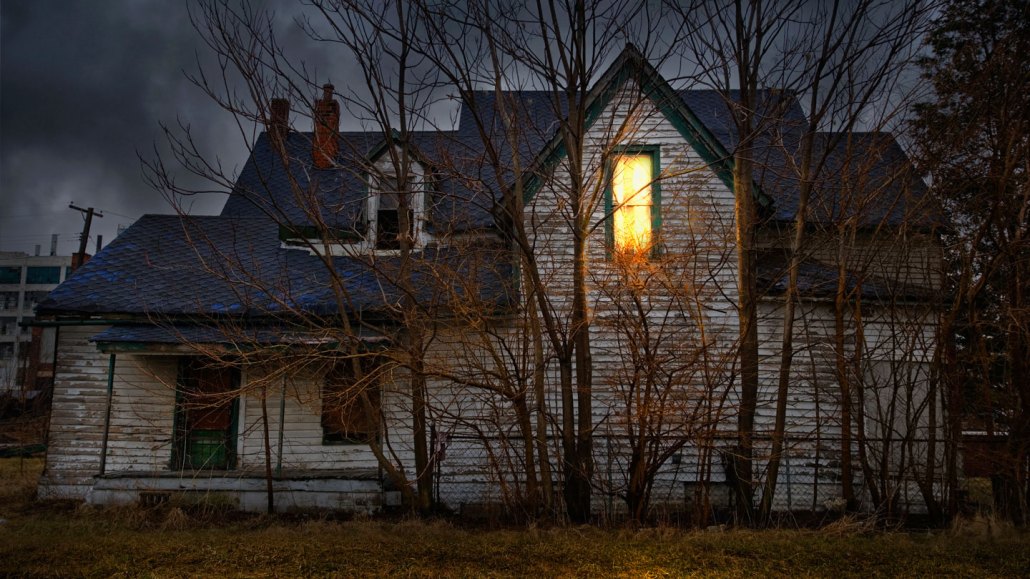 an old spooky house with one light on upstairs and surrounded by bare trees