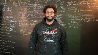Ronald Gamble stands in front of a blackboard covered with formulae. He's a black man with brown hair, a moustage and beard. He's wearing glasses and a sweatshirt that reads "NASA Goddard."