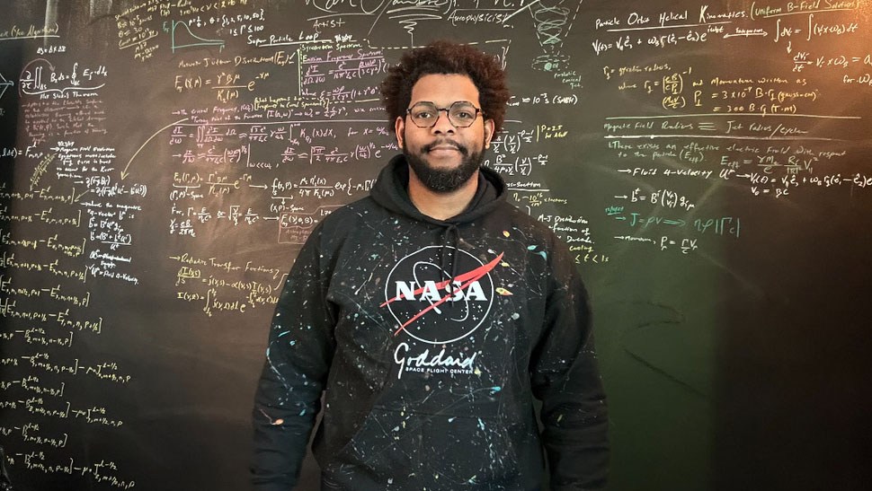 Ronald Gamble stands in front of a blackboard covered with formulae. He's a black man with brown hair, a moustage and beard. He's wearing glasses and a sweatshirt that reads "NASA Goddard."