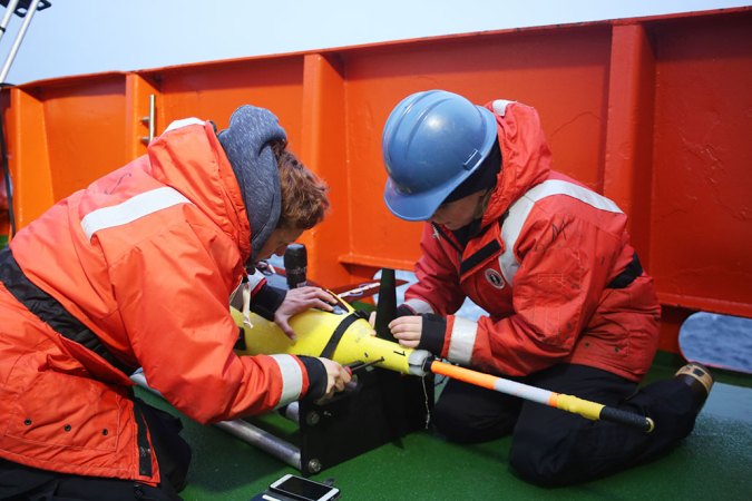 Two people wearing orange coats and blue construction hats tinker with a yellow sea glider.