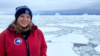 Oceanographer Lily Dove stands in front of pack ice in Antarctica. She is a white woman with blue eyes and blonde shoulder length hair. She is wearing a red coat and blue hat.