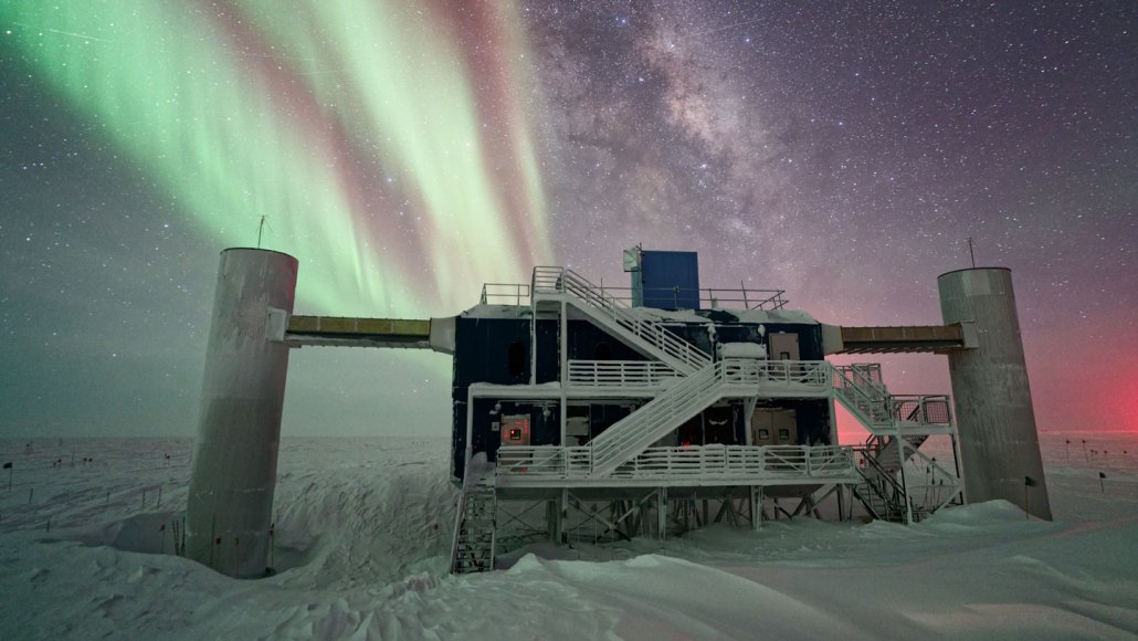 Beneath a brilliant aurora in a night sky, a building stands amid a field of snow and ice.
