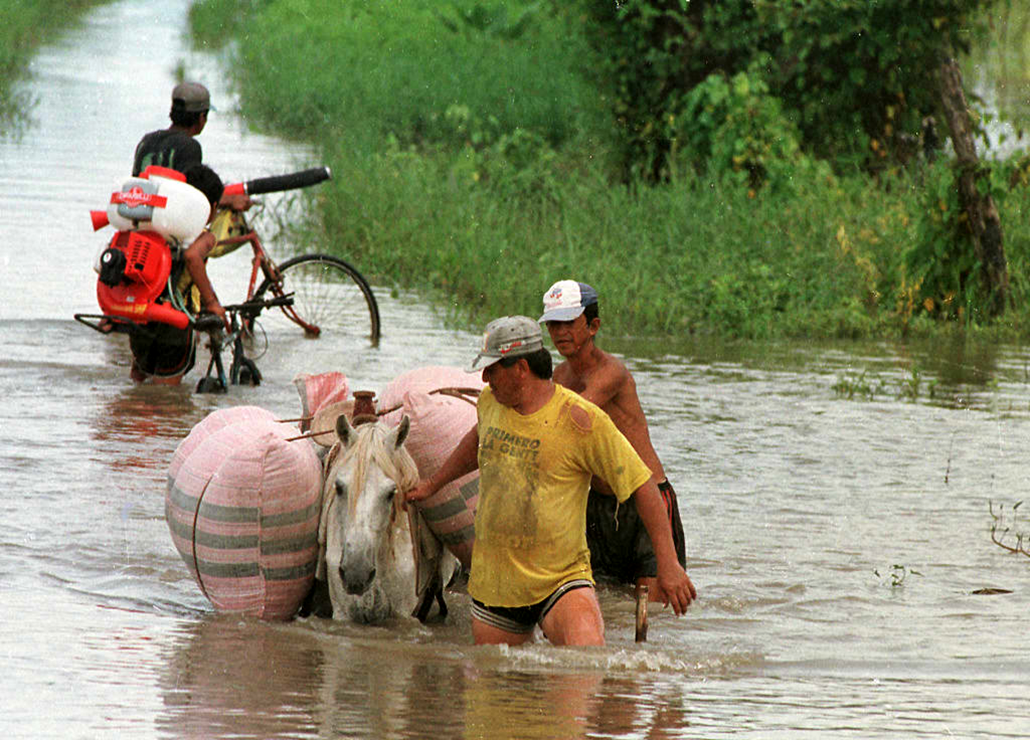 A group of men pulling a horse burdened with bags through flood water, behind them another man on a bicycle walks his bike through the water