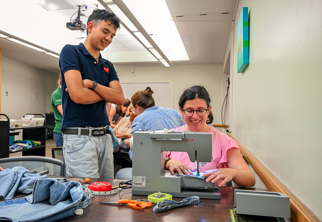 a woman sewing blue jean material at a sewing machine as a helper looks on