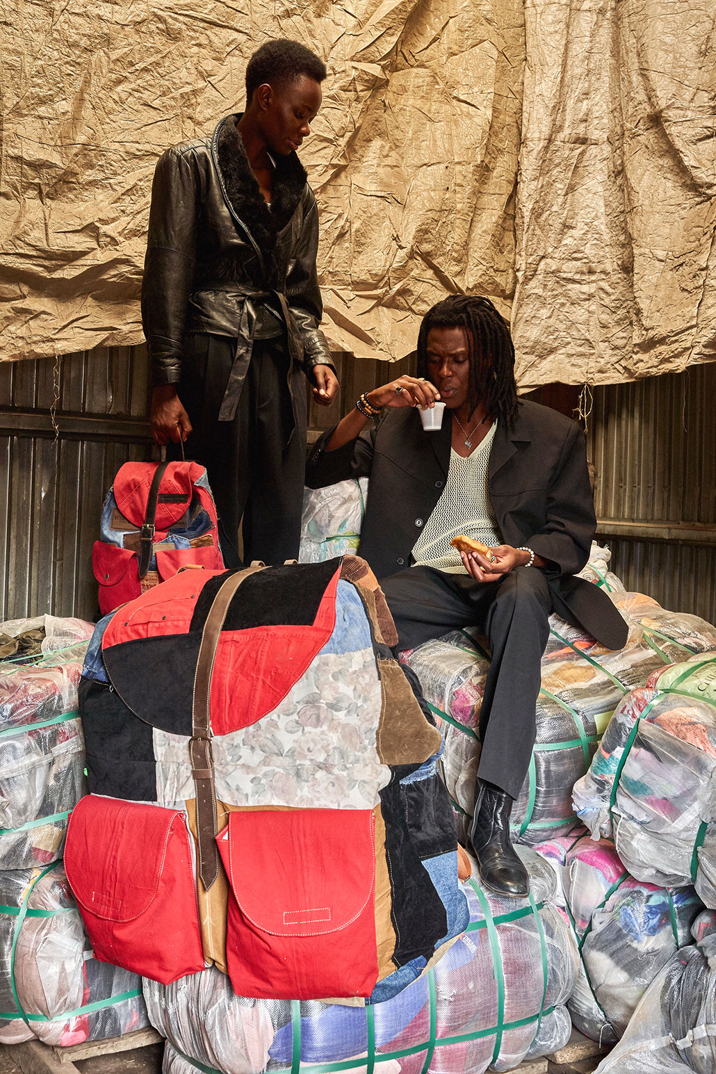 a photo of two models sitting on bales of recycled clothing. They both have a designer backpack made from denim, cordoroy, leather and other recycled fabrics.