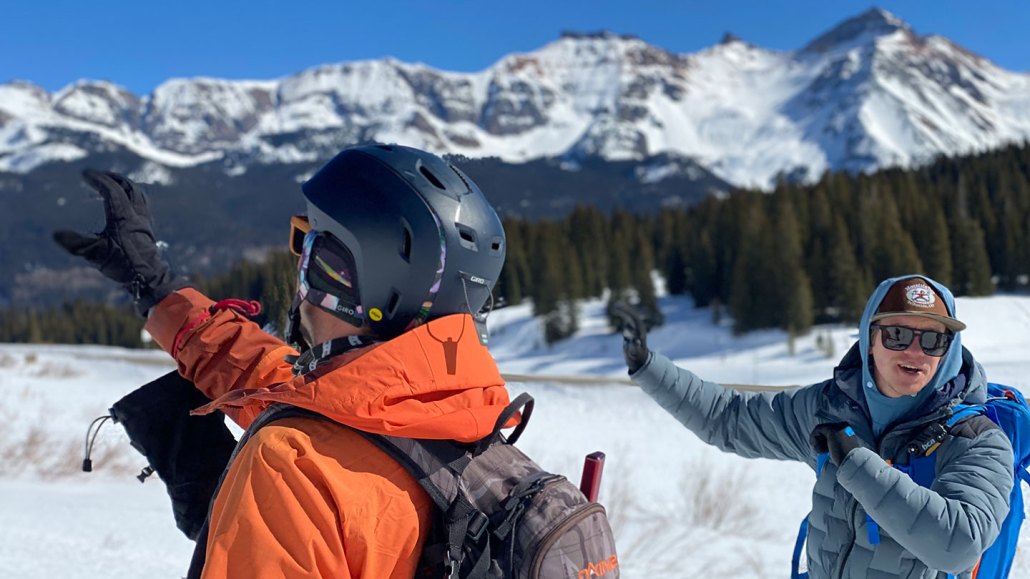 Avalanche forecaster Chris Dickson stands next to a person wearing an orange winter jacket and winter gear. Dickson is a white man with blond hair. He is wearing a gray winter jacket and sunglasses. His hands are raised as he talks to someone behind the camera.