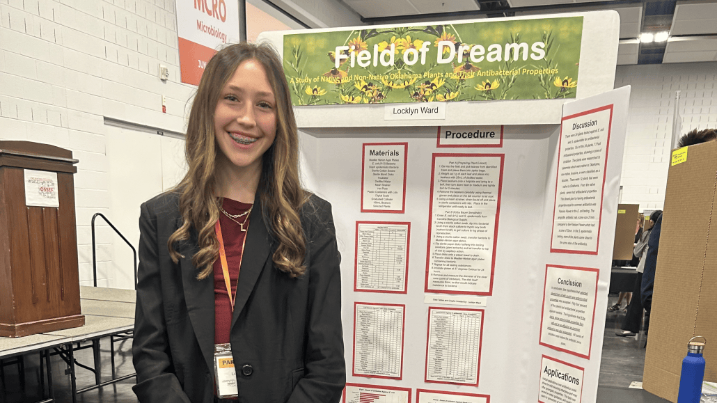 a teen girl smiles in front of a project poster titled 'field of dreams' on a background of flowering plants